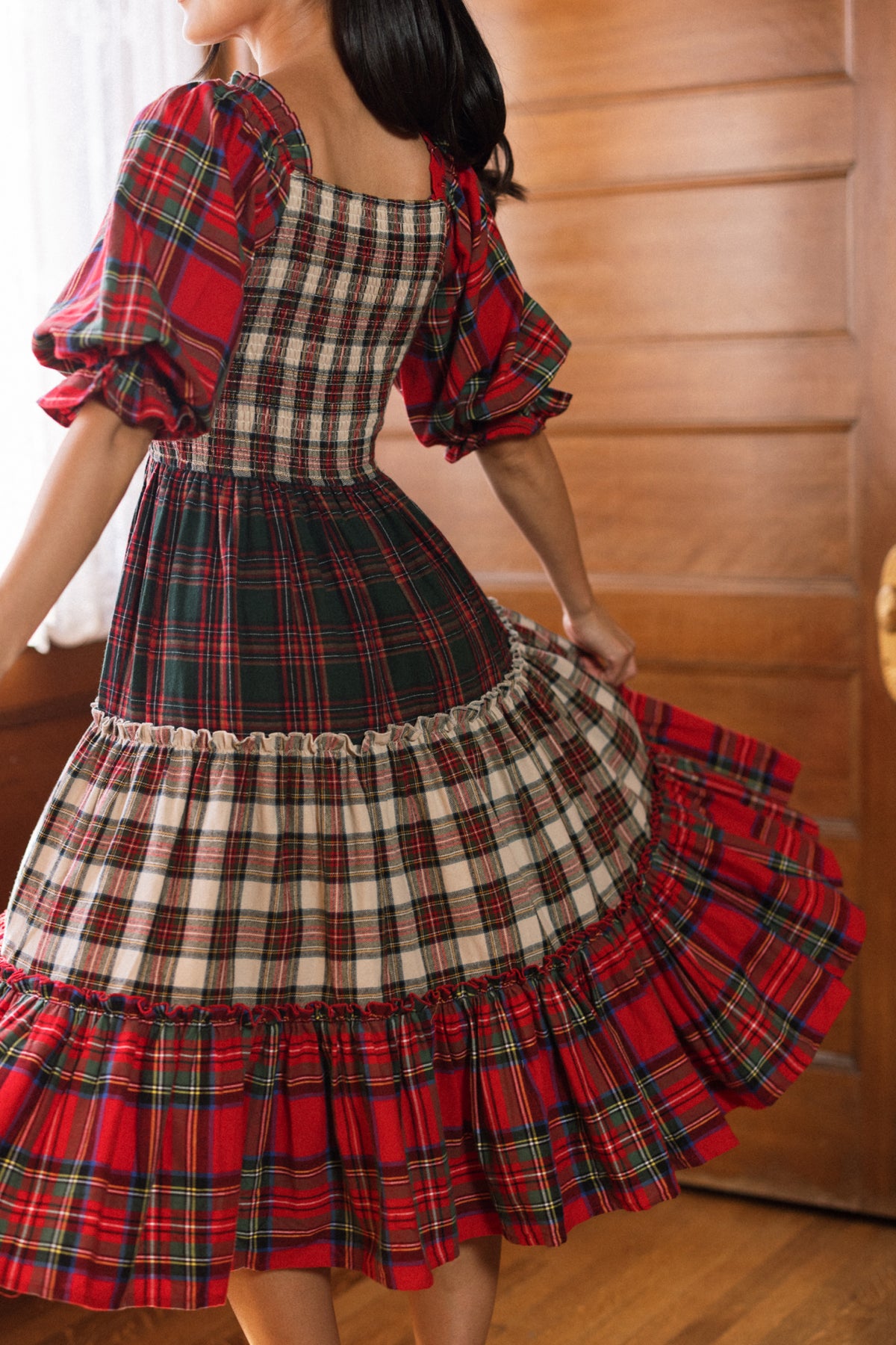 Wearing the Ivy City Co Madeline Dress in Mixed Plaid, a person twirls indoors, revealing the movement of its flowing skirt and puffed sleeves. The warm wooden backdrop and soft lighting add to the cozy atmosphere.
