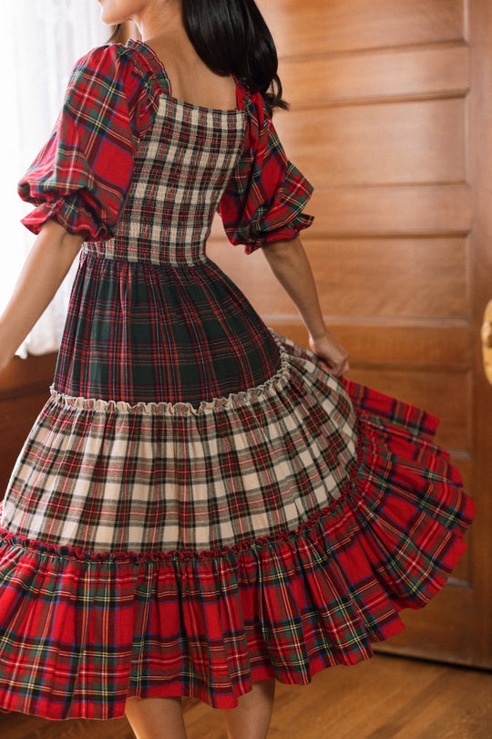 Wearing the Ivy City Co Madeline Dress in Mixed Plaid, a person twirls indoors, revealing the movement of its flowing skirt and puffed sleeves. The warm wooden backdrop and soft lighting add to the cozy atmosphere.