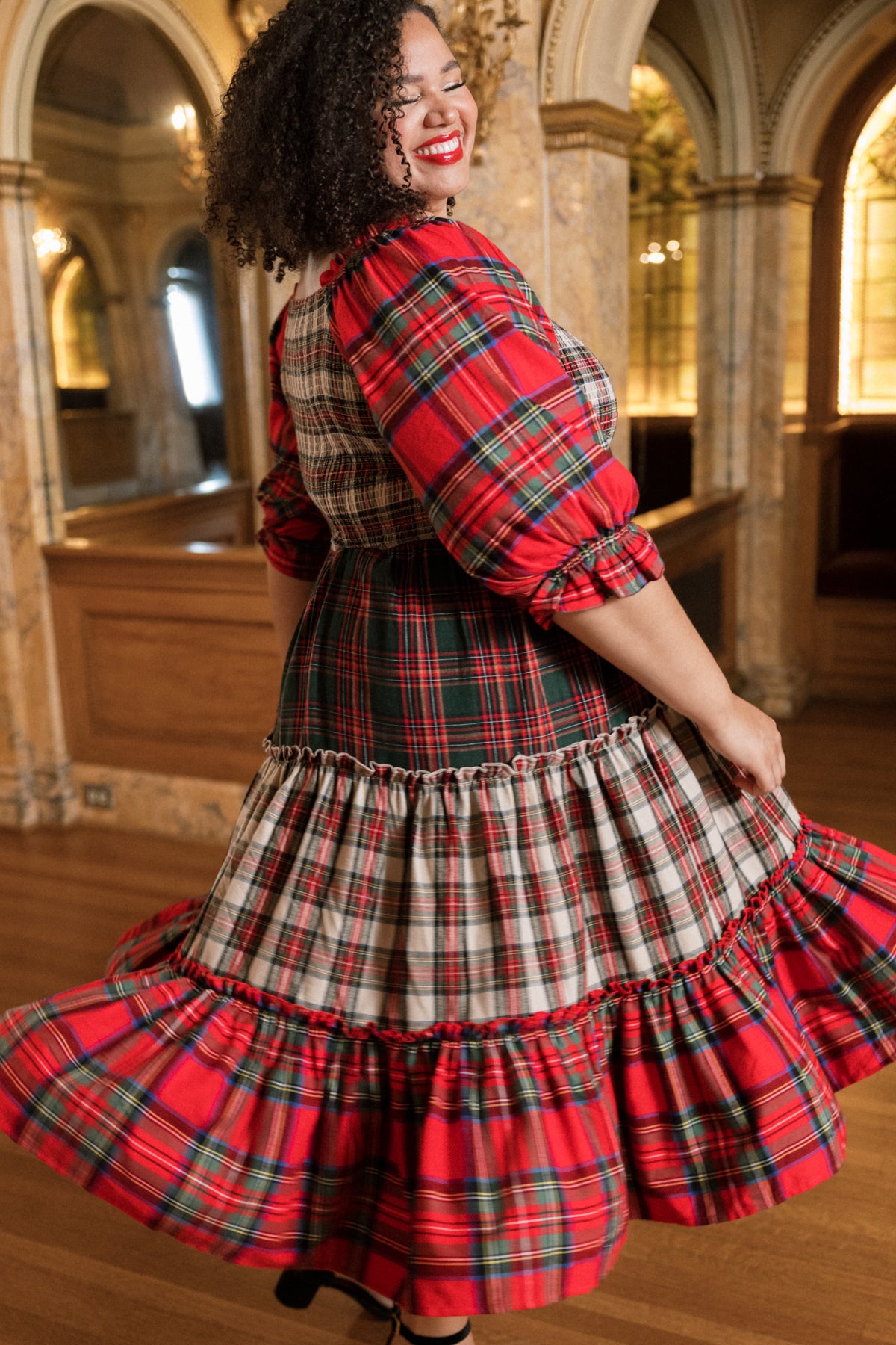 A woman with curly hair twirls in the Ivy City Co Madeline Dress in Mixed Plaid—a flowing, red plaid dress—inside an elegant, ornate room with arched mirrors and wooden floors, making it the perfect choice for a cozy Christmas family dress.