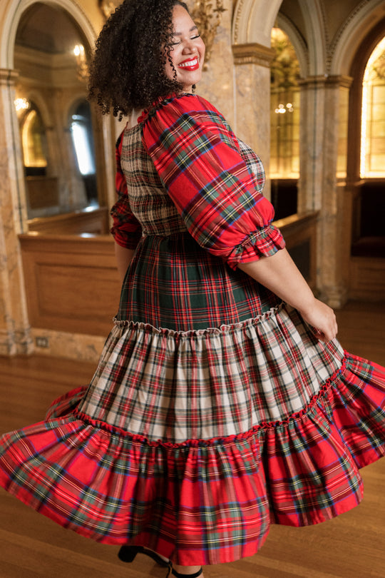 A woman with curly hair twirls in the Ivy City Co Madeline Dress in Mixed Plaid—a flowing, red plaid dress—inside an elegant, ornate room with arched mirrors and wooden floors, making it the perfect choice for a cozy Christmas family dress.