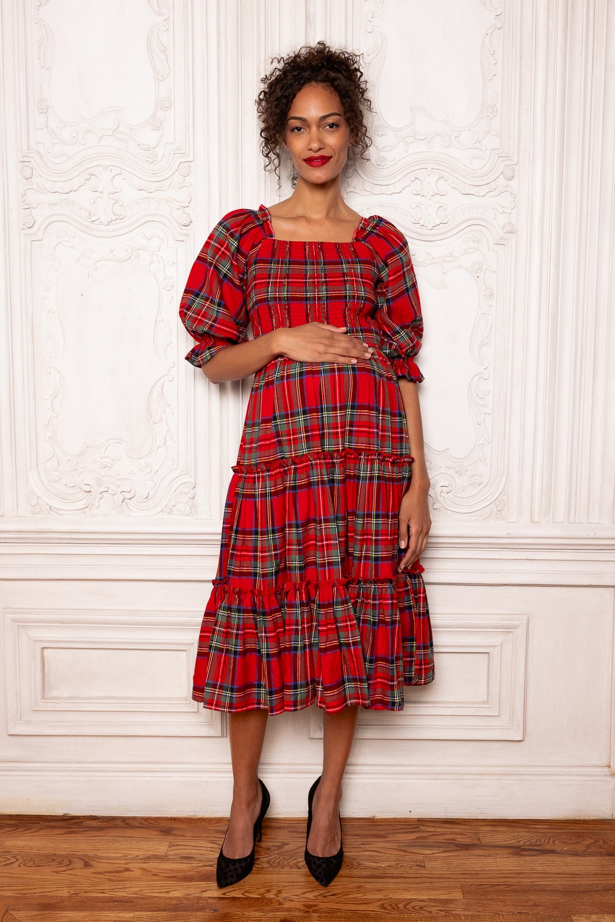 A woman stands on a wooden floor against an ornate white wall, smiling with one hand gently resting on her stomach—showcasing the maternity-friendly design of the Ivy City Co Madeline Dress in Holiday Plaid, which features puff sleeves and a tiered midi silhouette.