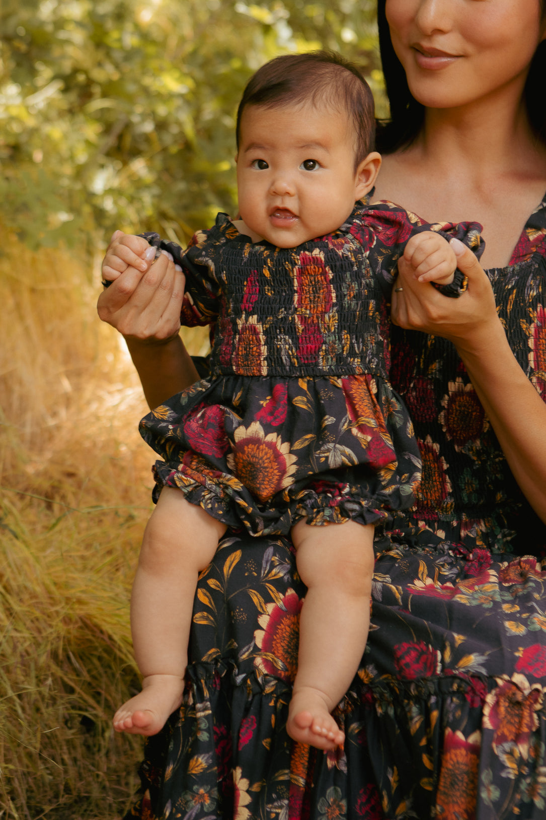 Woman in a floral dress holding a baby in a similar dress outdoors.