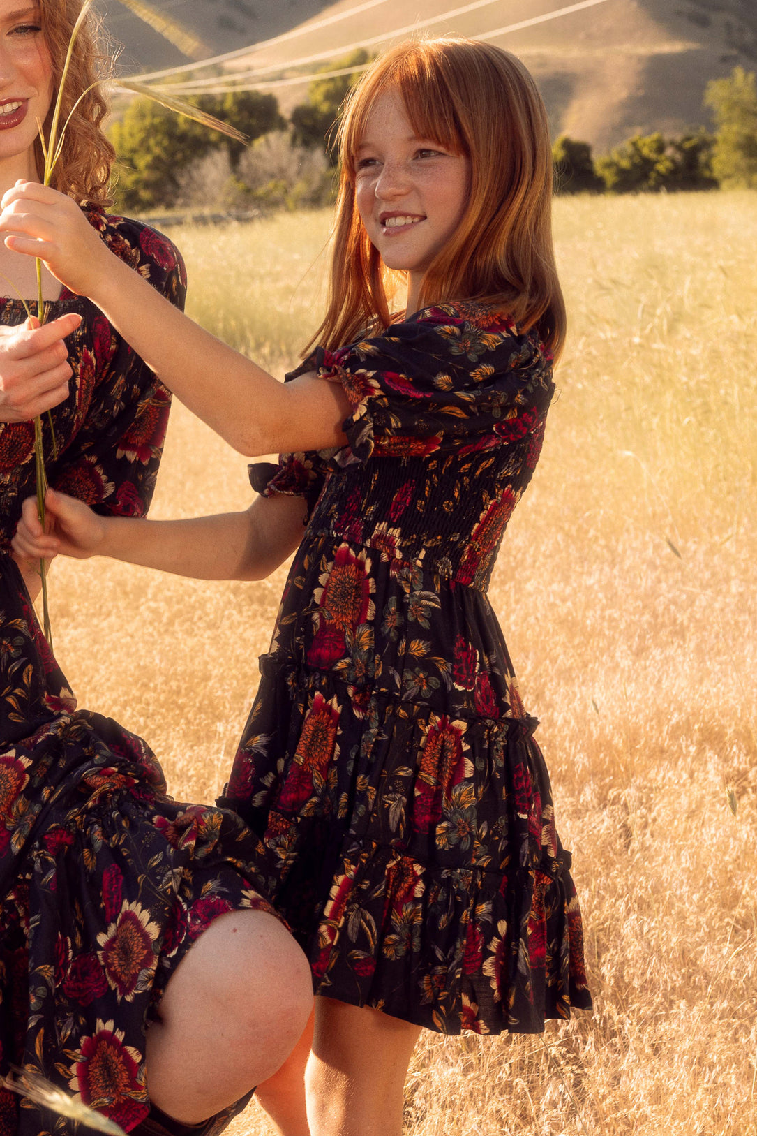Two young girls in patterned dresses standing in a field with mountains in the background.