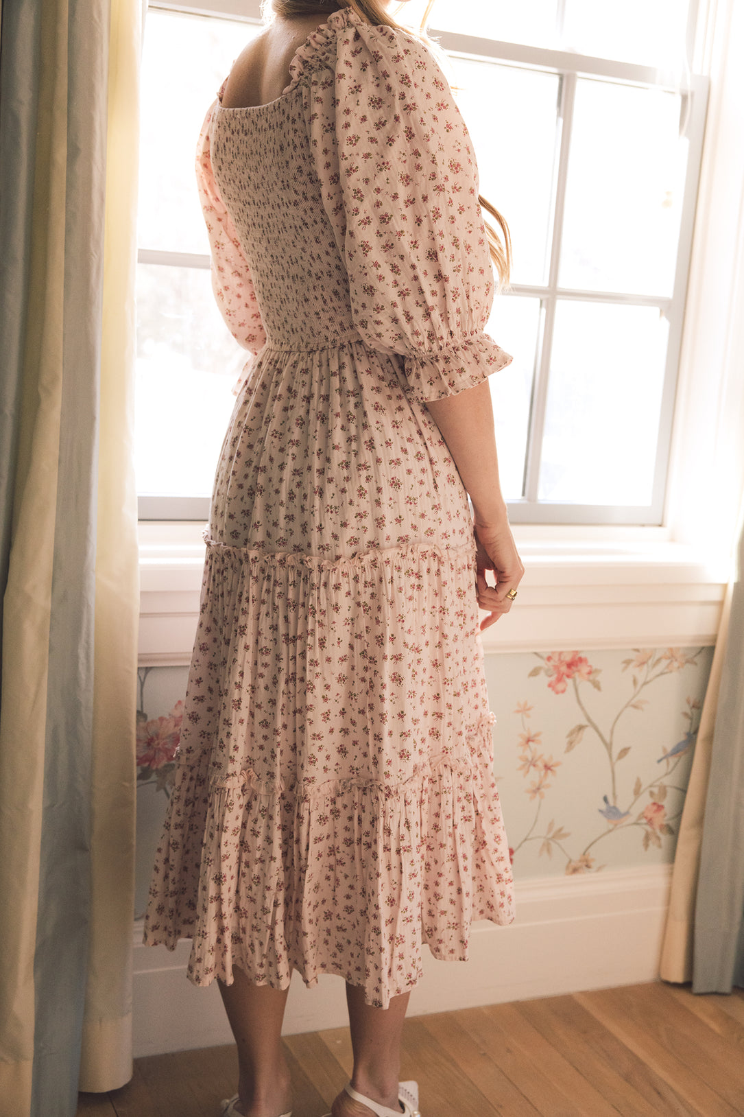 A woman stands indoors by a window, shown from the back in Ivy City Co's Madeline Dress In Pink—a light pink floral maternity-friendly dress with long sleeves, a smocked bodice, and tiered skirt as sunlight streams into the room.