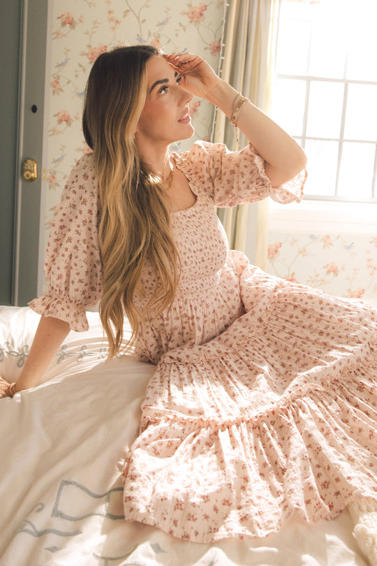 A woman with long blonde hair wears the Ivy City Co Madeline Dress in Pink, sitting thoughtfully on a sunlit bed with floral wallpaper. This elegant dress is both stylish and maternity friendly.