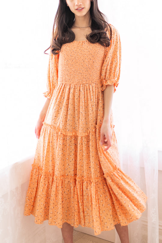 A woman wears the Madeline Dress in Sorbet by Ivy City Co.—a peach, floral maternity dress with a smocked bodice and puff sleeves—indoors, holding her skirt. The bright background highlights its nursing-friendly design.