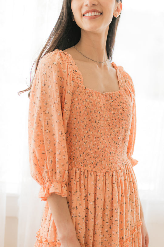 A woman smiles in the Ivy City Co. Madeline Dress in Sorbet, a peach maternity dress with puffed sleeves, a smocked bodice, and a small floral print. The softly lit, blurred background enhances the gentle mood.