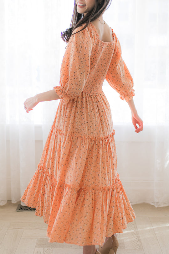 A woman twirls gracefully indoors wearing the Ivy City Co. Madeline Dress in Sorbet—a peach, long-sleeve floral maternity midi dress with a smocked bodice—her wavy hair flowing as natural light streams through sheer white curtains.