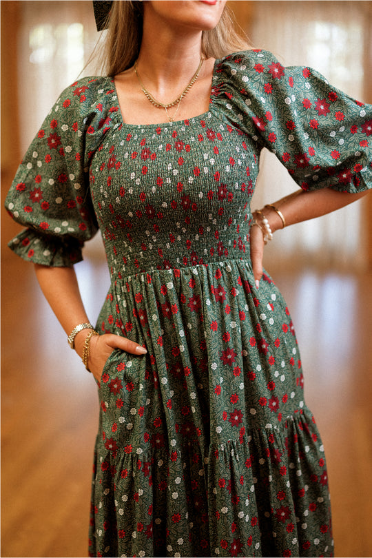 A woman stands indoors with her hands on her hips, wearing the Ivy City Co Madeline Maxi Dress in Evergreen Merry Ditsy. This elegant cotton dress features a smocked bodice, puffed sleeves, and a square neckline adorned with red and white floral prints. Designed to blend effortless style with comfort, it is both maternity and nursing friendly. She completes her look with gold jewelry.