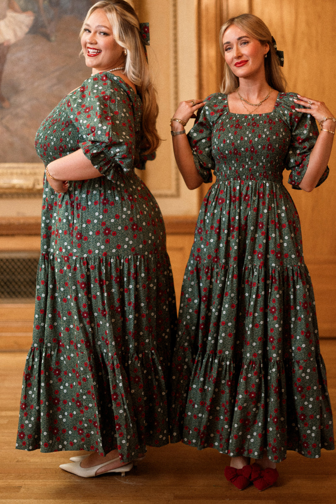 Two women stand indoors, smiling and posing in matching Madeline Maxi Dresses in Evergreen Merry Ditsy by Ivy City Co. Designed with smocked cotton for a comfortable fit that’s both maternity and nursing friendly, one woman faces sideways while the other stands forward. Both have long blonde hair and are in a warmly lit room with wooden walls.