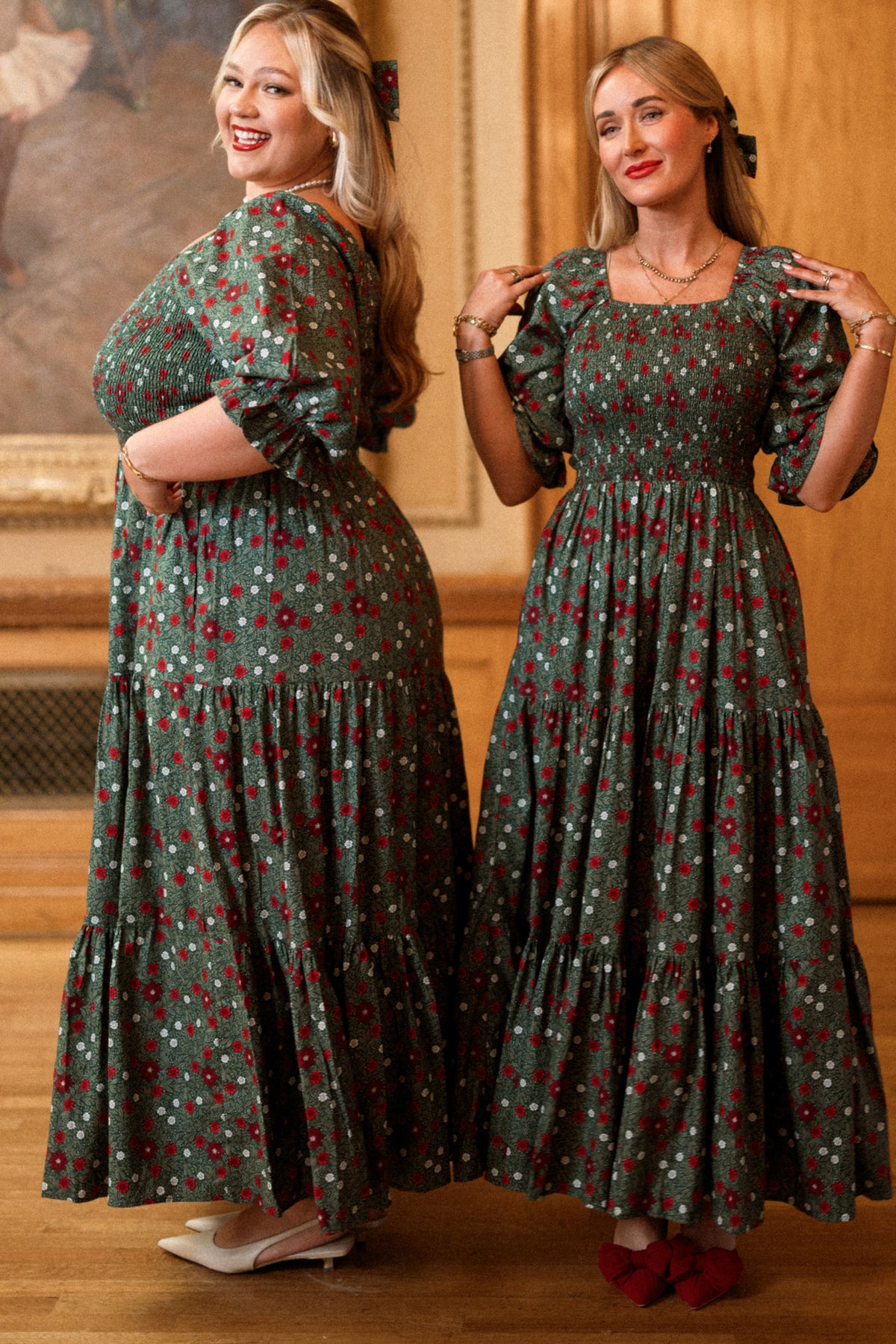 Two women stand indoors, smiling and posing in matching Madeline Maxi Dresses in Evergreen Merry Ditsy by Ivy City Co. Designed with smocked cotton for a comfortable fit that’s both maternity and nursing friendly, one woman faces sideways while the other stands forward. Both have long blonde hair and are in a warmly lit room with wooden walls.