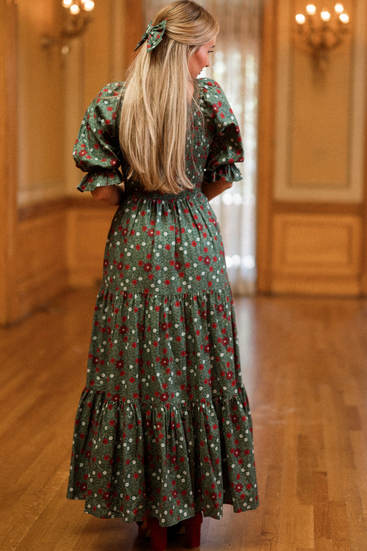 A woman with long blonde hair stands indoors on a wooden floor, facing away from the camera. She wears the Madeline Maxi Dress in Evergreen Merry Ditsy by Ivy City Co., its smocked cotton design providing both maternity and nursing-friendly comfort. Ornate wood paneling and chandeliers create an elegant backdrop for this charming floral dress.