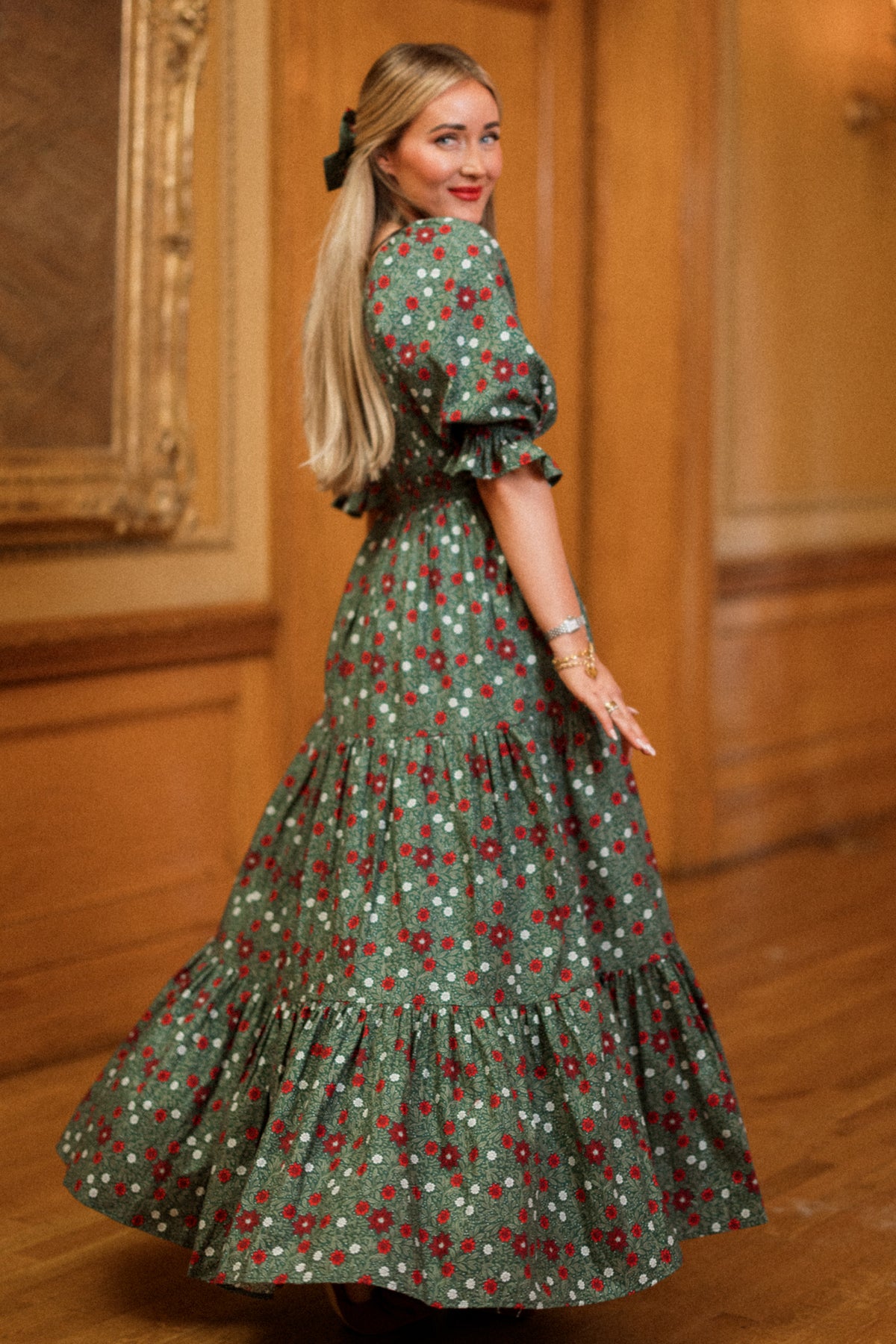 A woman with long blonde hair twirls and smiles in an ornate room with wooden floors and a framed painting on the wall, wearing the Ivy City Co Madeline Maxi Dress in Evergreen Merry Ditsy. This smocked cotton dress is both maternity and nursing friendly, combining comfort and style for a timeless look.