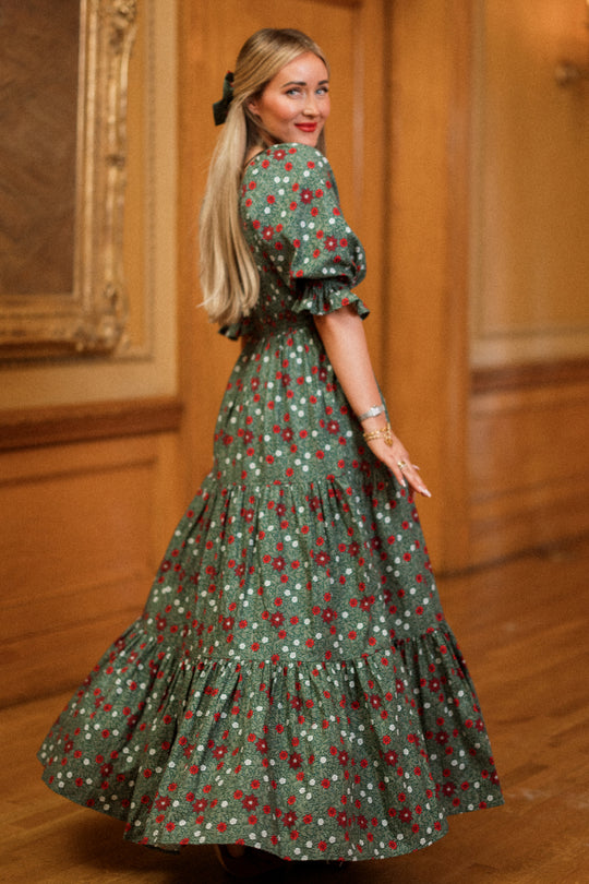 A woman with long blonde hair twirls and smiles in an ornate room with wooden floors and a framed painting on the wall, wearing the Ivy City Co Madeline Maxi Dress in Evergreen Merry Ditsy. This smocked cotton dress is both maternity and nursing friendly, combining comfort and style for a timeless look.
