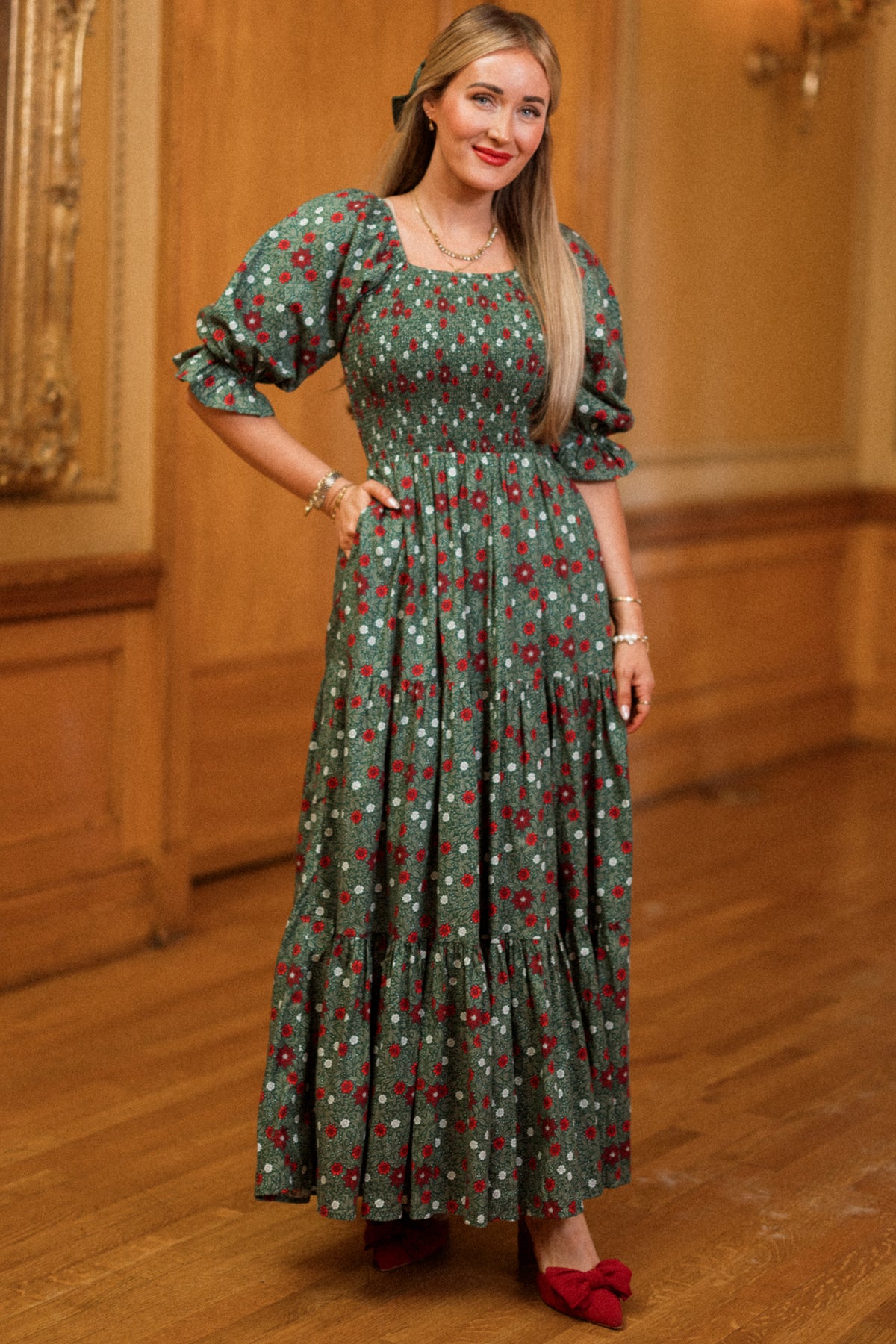 A woman stands indoors, smiling with one hand in her pocket, wearing the Ivy City Co Madeline Maxi Dress in Evergreen Merry Ditsy—a long floral dress featuring puffed sleeves and a smocked cotton design that’s both maternity and nursing friendly. She pairs it with red shoes, her long straight hair complementing the stylish yet practical look amid wooden paneling and ornate decor.