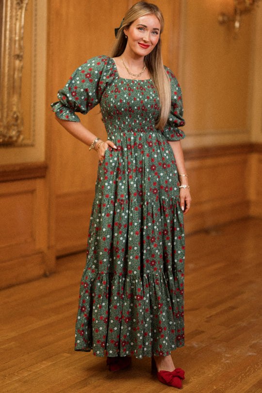 A woman stands indoors, smiling with one hand in her pocket, wearing the Ivy City Co Madeline Maxi Dress in Evergreen Merry Ditsy—a long floral dress featuring puffed sleeves and a smocked cotton design that’s both maternity and nursing friendly. She pairs it with red shoes, her long straight hair complementing the stylish yet practical look amid wooden paneling and ornate decor.