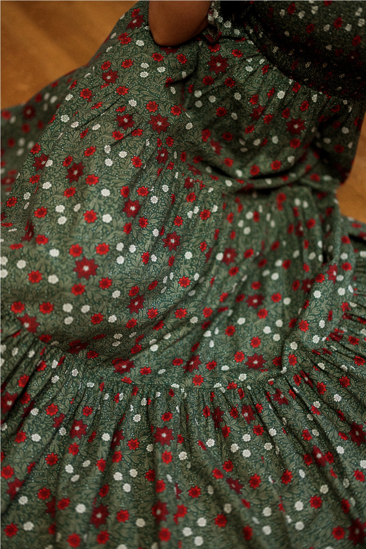 A close-up of the Madeline Maxi Dress in Evergreen Merry Ditsy by Ivy City Co reveals a delicate pattern of small red and white flowers. The soft, textured smocked cotton fabric provides a comfortable fit, making this dress both maternity and nursing friendly, while a hand gently gathers the skirt to showcase its detail.