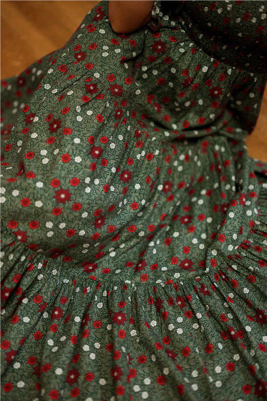A close-up of the Madeline Maxi Dress in Evergreen Merry Ditsy by Ivy City Co reveals a delicate pattern of small red and white flowers. The soft, textured smocked cotton fabric provides a comfortable fit, making this dress both maternity and nursing friendly, while a hand gently gathers the skirt to showcase its detail.