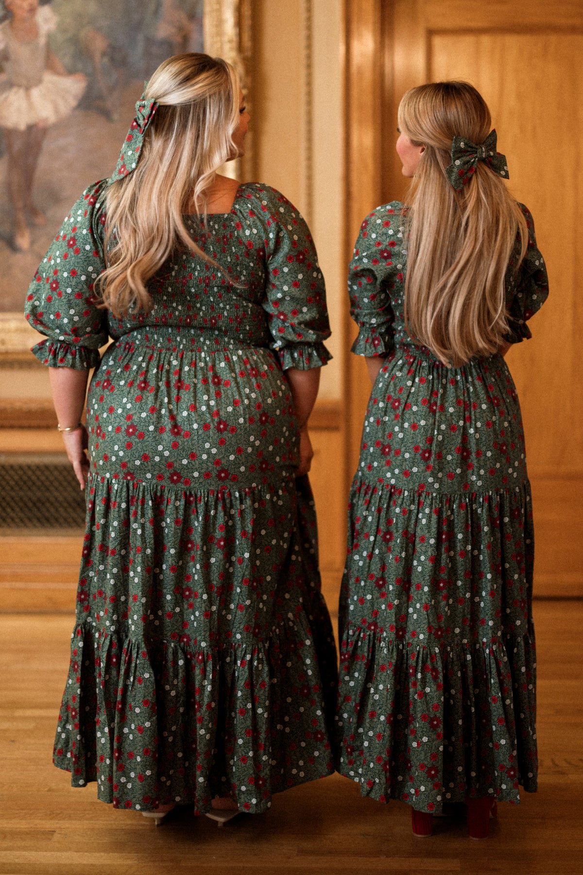 Two women with long blond hair, dressed in matching Ivy City Co Madeline Maxi Dresses in Evergreen Merry Ditsy, stand side by side indoors, facing away from the camera in a warmly lit room with wooden paneling and art on the wall. Their smocked cotton dresses are both maternity and nursing friendly, making them as practical as they are stylish.