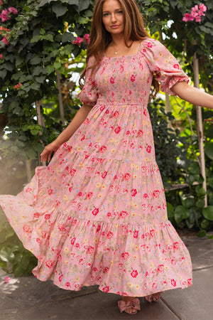 A woman with long hair wears the Ivy City Co Madeline Maxi Dress in Pink Vintage Poppies, a floral puff-sleeve, tiered maxi that's maternity friendly, standing outdoors among lush plants and flowers while holding out her flowing dress.