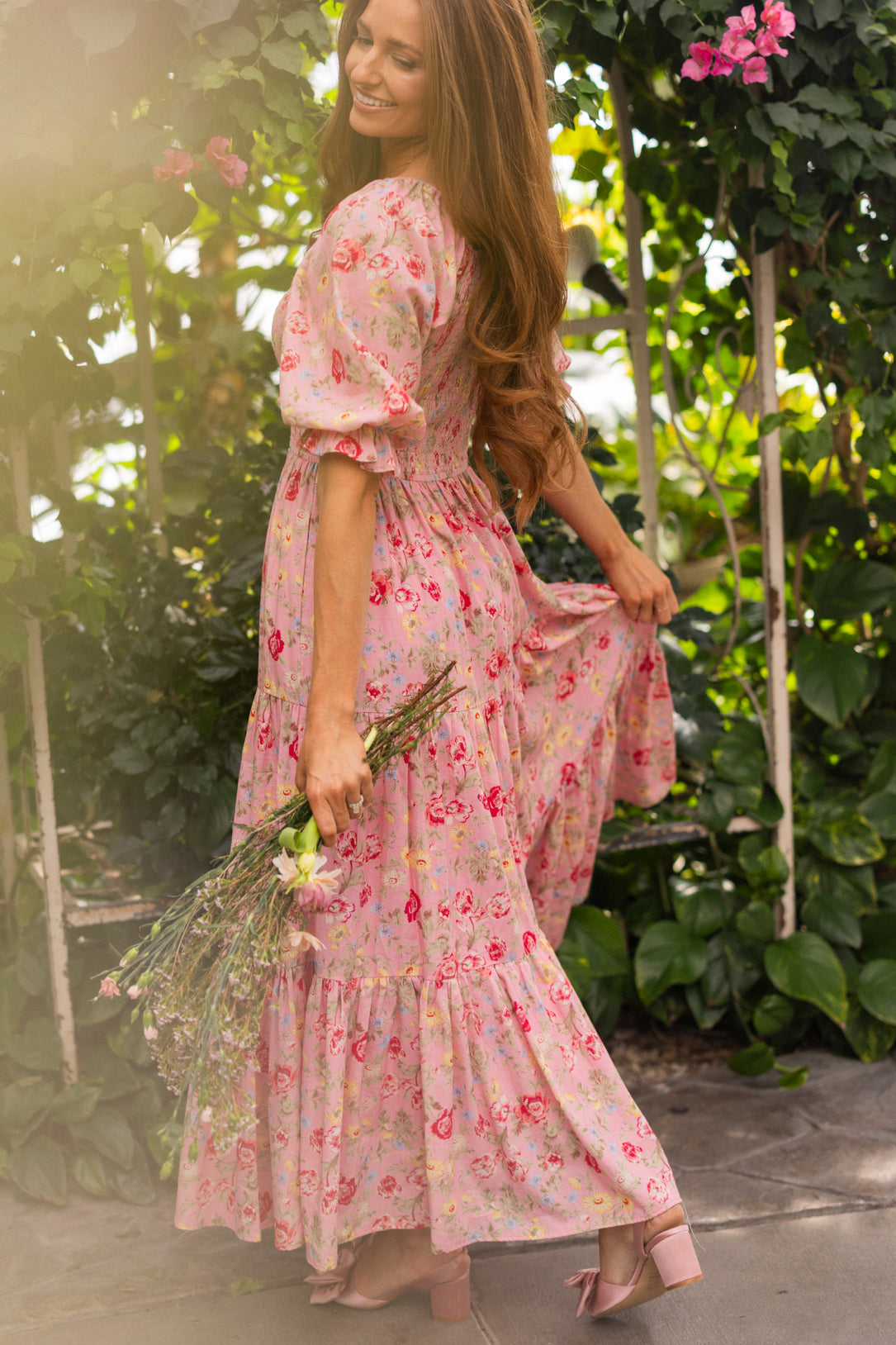 A woman in the Ivy City Co Madeline Maxi Dress in Pink Vintage Poppies stands outdoors, smiling over her shoulder with a wildflower bouquet, surrounded by lush greenery and blooming pink flowers.