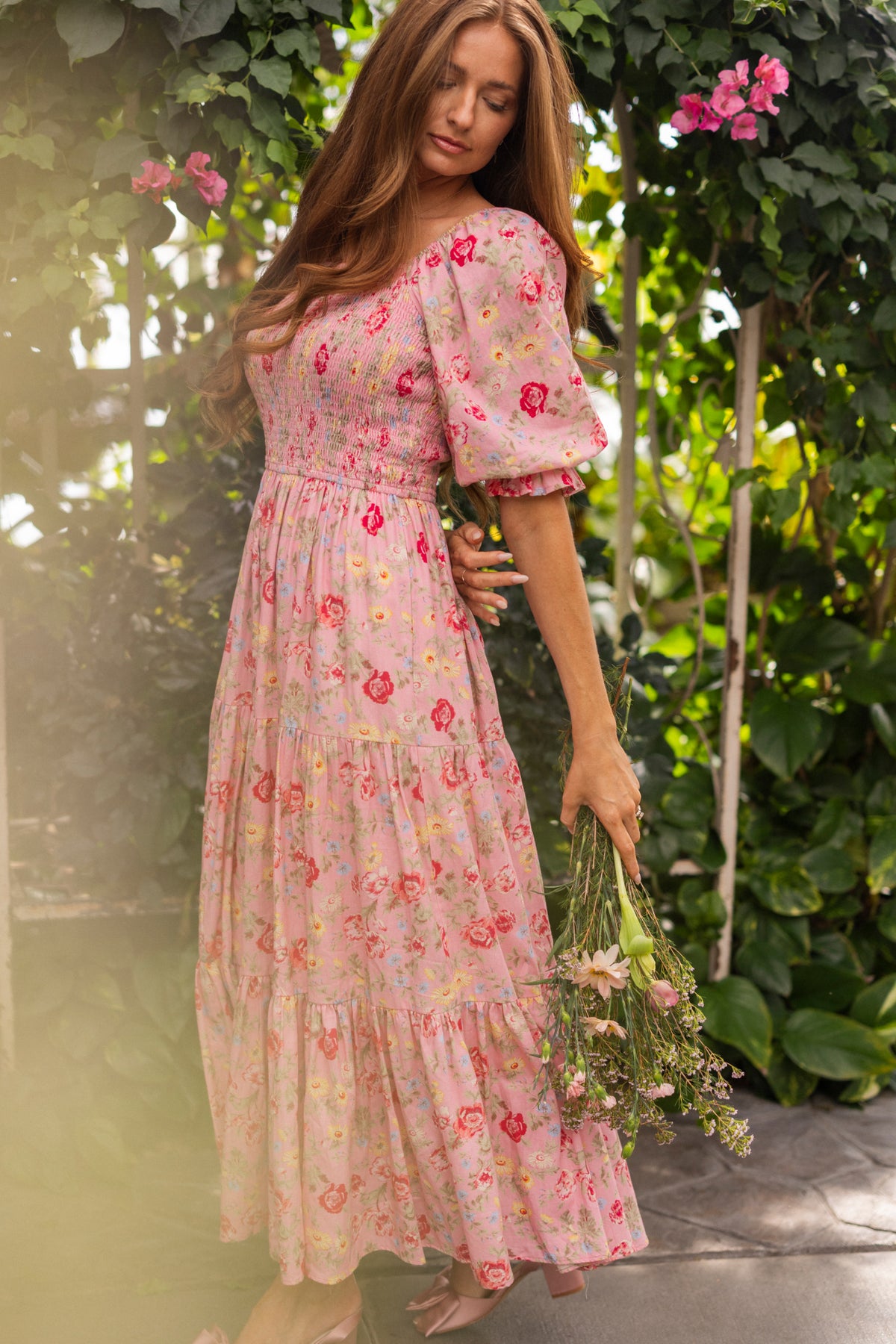 A woman in the Ivy City Co Madeline Maxi Dress in Pink Vintage Poppies stands barefoot in a lush garden, holding wildflowers amid green leaves and pink blossoms. This elegant maxi is also maternity friendly.