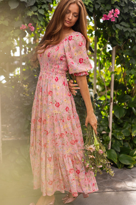 A woman in the Ivy City Co Madeline Maxi Dress in Pink Vintage Poppies stands barefoot in a lush garden, holding wildflowers amid green leaves and pink blossoms. This elegant maxi is also maternity friendly.