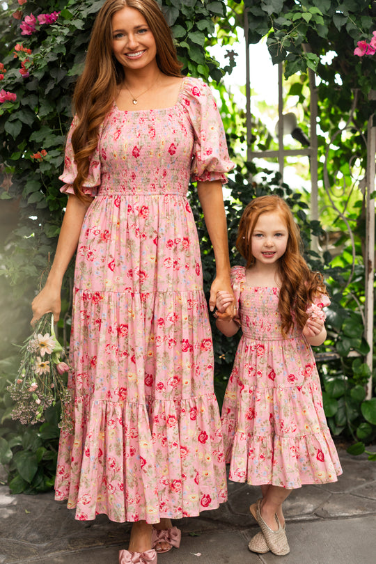 A woman and a young girl smile and hold hands in matching Ivy City Co Madeline Maxi Dress in Pink Vintage Poppies, surrounded by lush greenery. This maternity-friendly dress is perfect for all occasions.