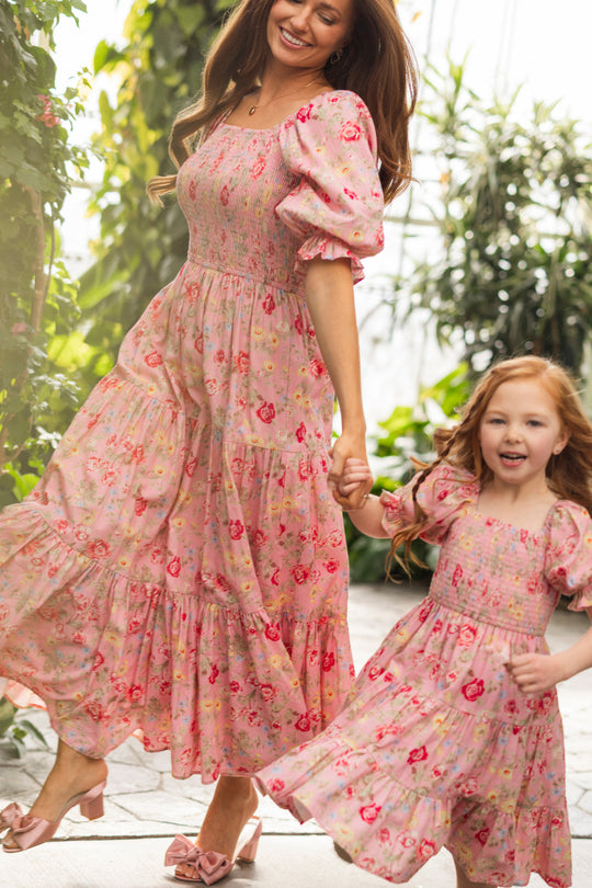 A woman and a young girl, both in matching Ivy City Co Madeline Maxi Dresses in Pink Vintage Poppies, smile and hold hands as they walk outdoors among lush greenery. This stylish floral maxi dress is comfortable and maternity friendly.