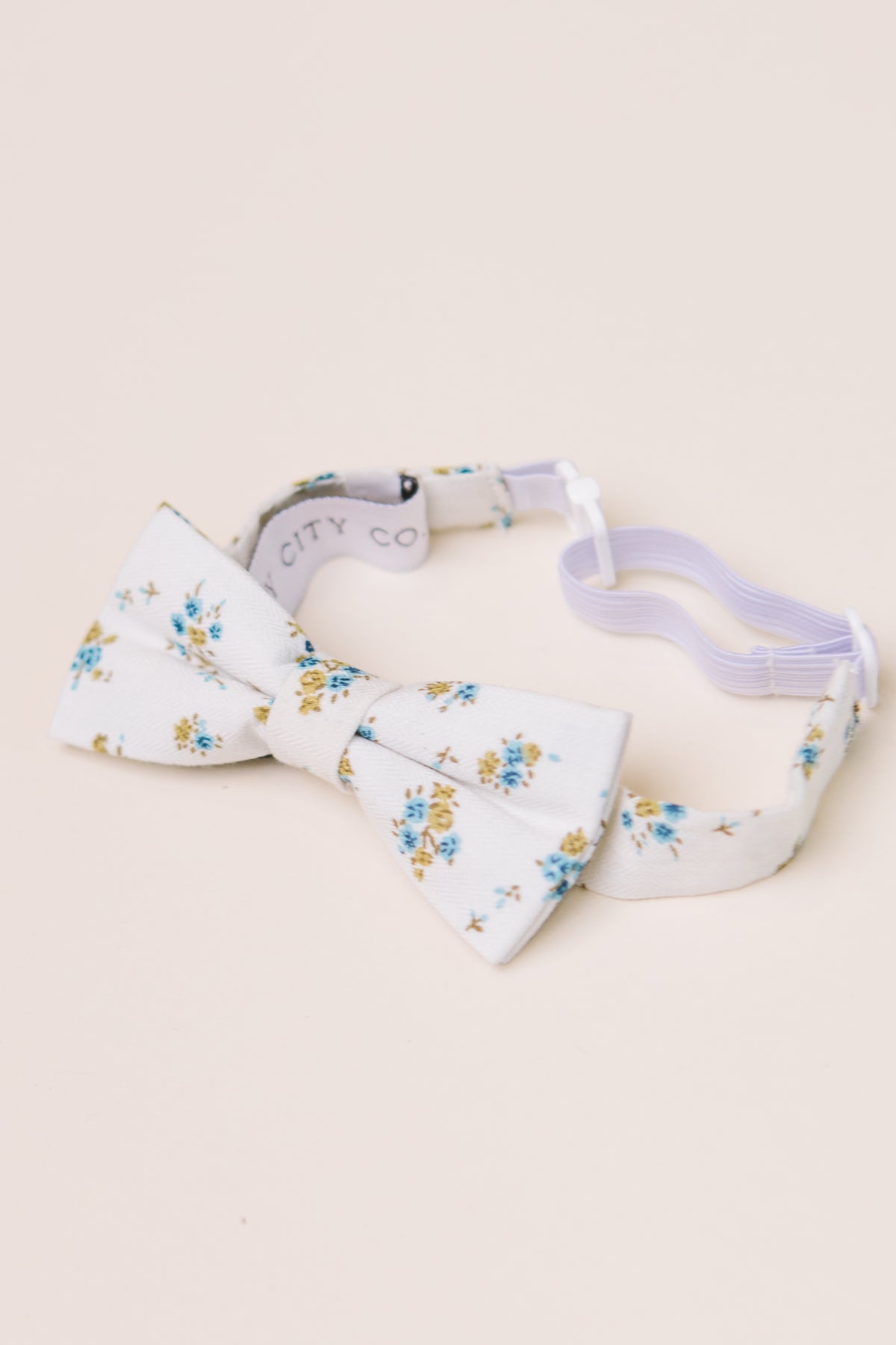 The Ivy City Co Ties in Madeline Blue Floral have a white base with blue and yellow blooms, an adjustable strap, and are great for family matching. Shown on a light beige background.