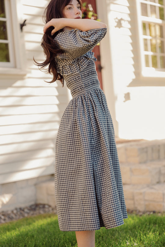A woman wearing the Mae 3/4 Sleeves Dress in Prussian Blue Plaid by Ivy City Co stands outside on green grass, one arm raised and her hair falling across her face, in front of a white house with stone accents as sunlight casts soft shadows.