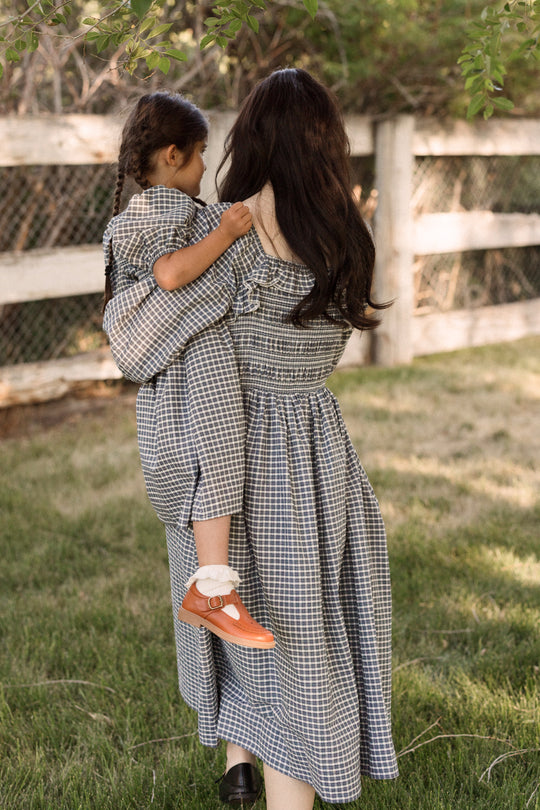 Certainly! Here’s a rewritten sentence incorporating your product data: A woman with long dark hair carries a young girl with braided hair, both dressed in matching Ivy City Co Mae 3/4 Sleeves Dresses in Prussian Blue Plaid. The smocked-bodice design makes these outfits comfortable and maternity friendly as they enjoy time outdoors near a wooden fence and trees.