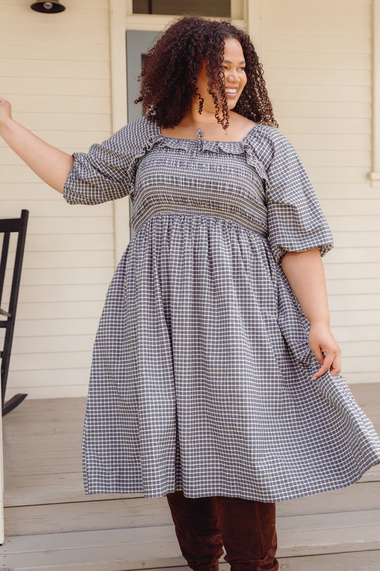 Smiling woman with curly hair wears the Ivy City Co Mae 3/4 Sleeves Dress in Prussian Blue Plaid, standing on a porch near a black rocking chair.