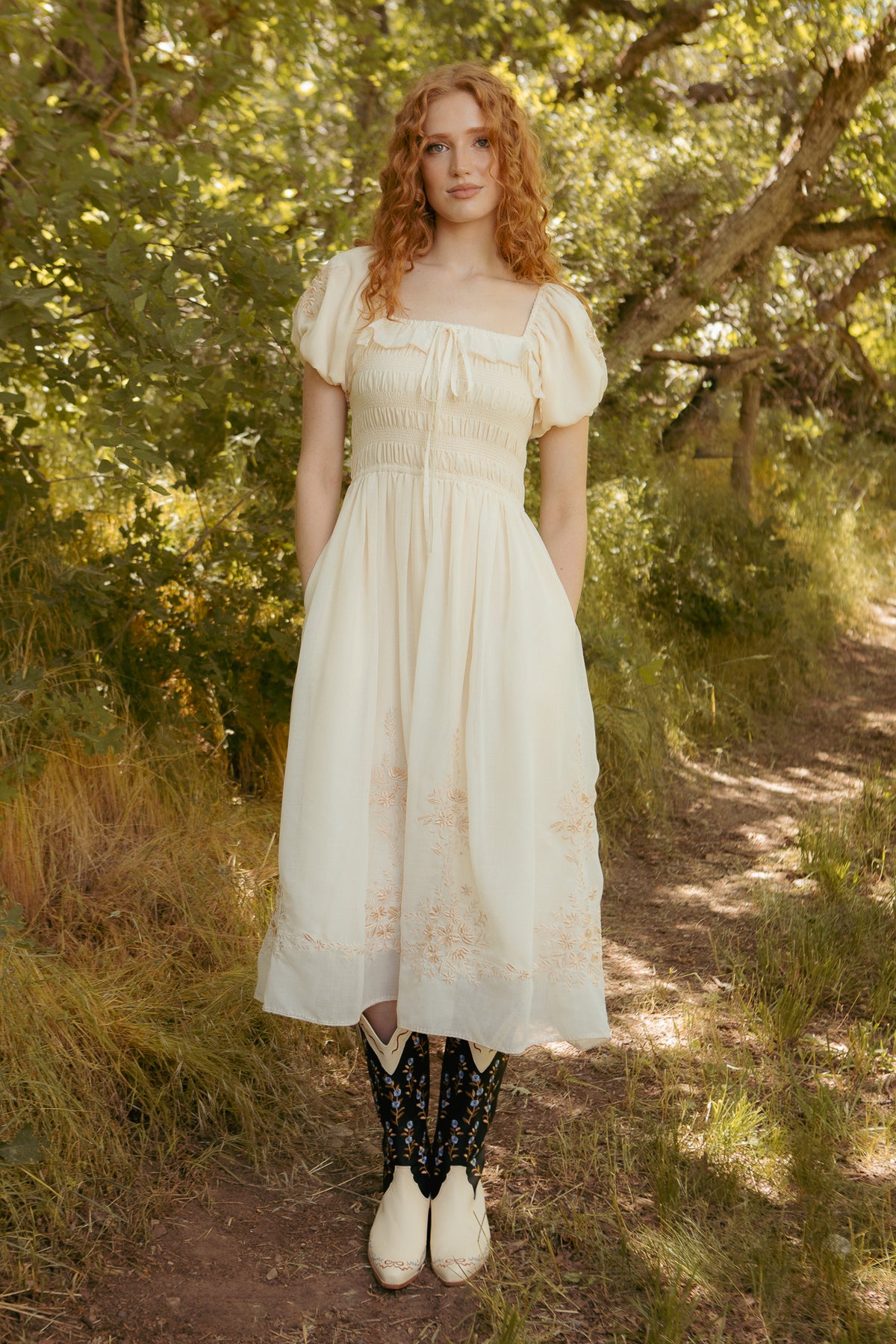 A woman with long red curly hair stands on a sunlit forest path, wearing the Mae Dress in Metallic Thread by Ivy City Co and white cowboy boots with black detailing.