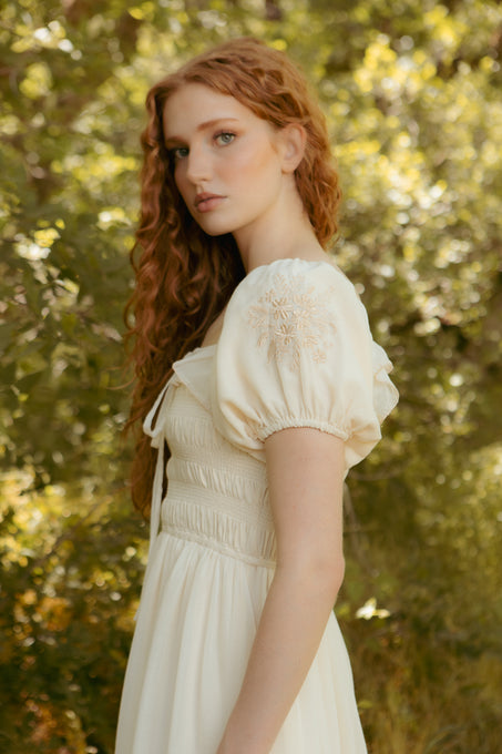 A woman with long, wavy red hair stands outdoors before leafy trees, wearing the Ivy City Co Mae Dress in Metallic Thread—a cream-colored, hand-embroidered floral dress with puffed sleeves—looking calmly at the camera.