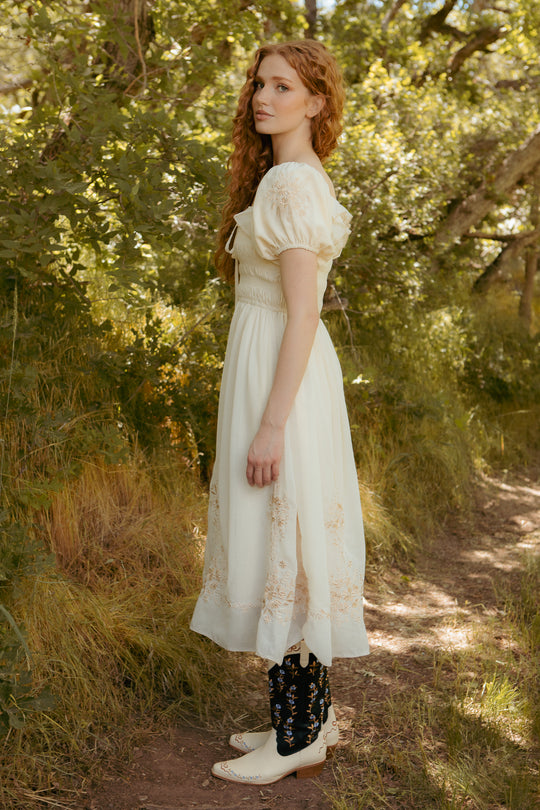 A woman with long curly red hair stands sideways on a sunlit forest path, wearing the Mae Dress in Metallic Thread by Ivy City Co, a cream-colored dress with hand-embroidered floral and lace details amidst lush green foliage.