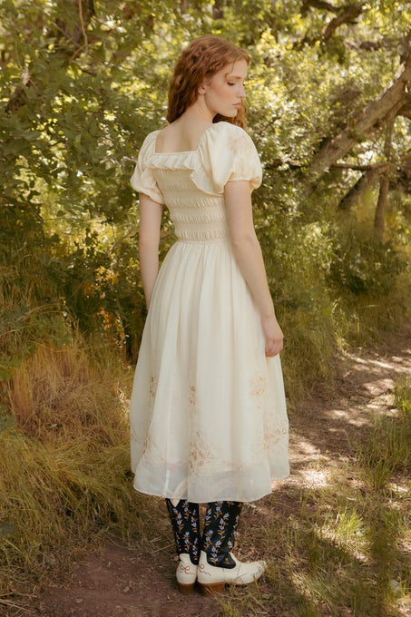 A woman with wavy red hair stands on a sunlit forest path, wearing the Mae Dress in Metallic Thread by Ivy City Co and black boots with blue and green patterns. She faces away, glancing to the side.