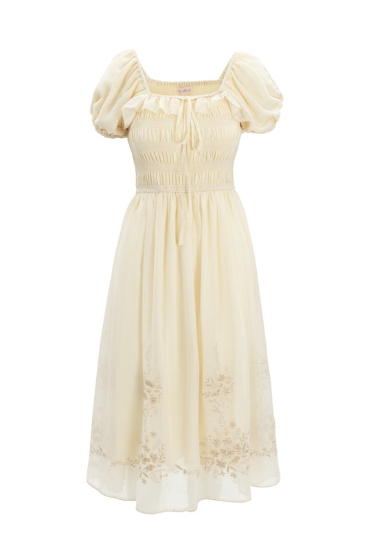 The Mae Dress in Metallic Thread by Ivy City Co is a cream, knee-length dress with hand-embroidered florals, short puff sleeves, a fitted smocked bodice, neckline tie, and subtle floral embroidery near the hem.