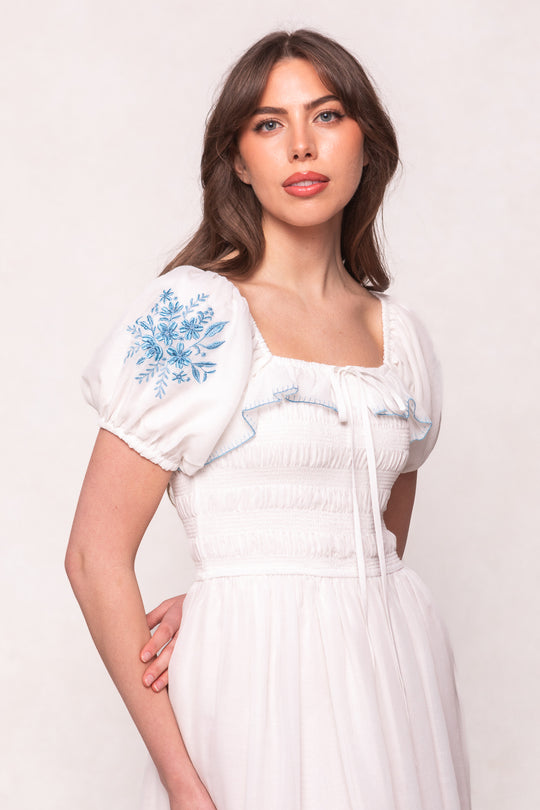 A woman with long brown hair wears the Ivy City Co Mae Dress in White with Blue Floral, featuring puff sleeves and a square neckline, standing against a light background.