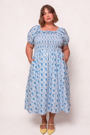 A woman with short, light brown hair wears the Ivy City Co Mae Dress in Blue Floral Stamp—nursing friendly with puff sleeves and pockets—paired with yellow pointed-toe flats, standing against a plain light background.