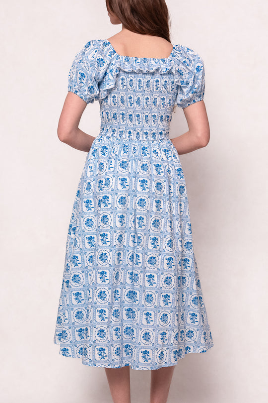 A woman faces away, wearing the Ivy City Co Mae Dress in Blue Floral Stamp. This nursing-friendly midi dress features puff sleeves, a smocked bodice, and a geometric tile pattern that falls below the knees.