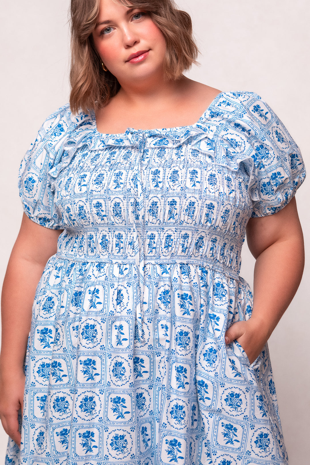 A woman with short brown hair wears the Ivy City Co Mae Dress in Blue Floral Stamp, featuring puffed sleeves, a smocked waist, nursing-friendly design, and pockets, standing against a light background.