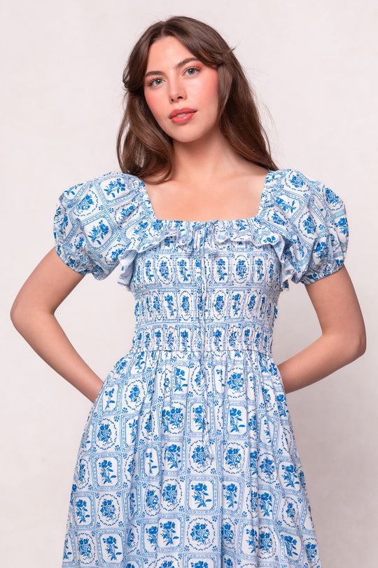 A woman with long brown hair models the Ivy City Co Mae Dress in Blue Floral Stamp, featuring puffed short sleeves and a square neckline. This nursing-friendly dress blends style and comfort effortlessly against a neutral backdrop.