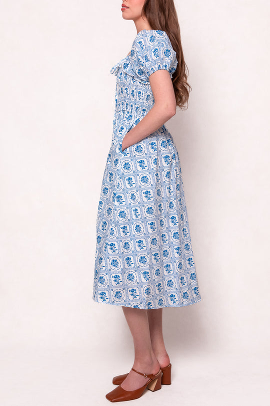 A woman stands sideways in the Mae Dress in Blue Floral Stamp by Ivy City Co, featuring short sleeves, pockets, and a midi length. She pairs it with brown low-heeled shoes; her long straight hair contrasts the plain, light background. Nursing friendly style.