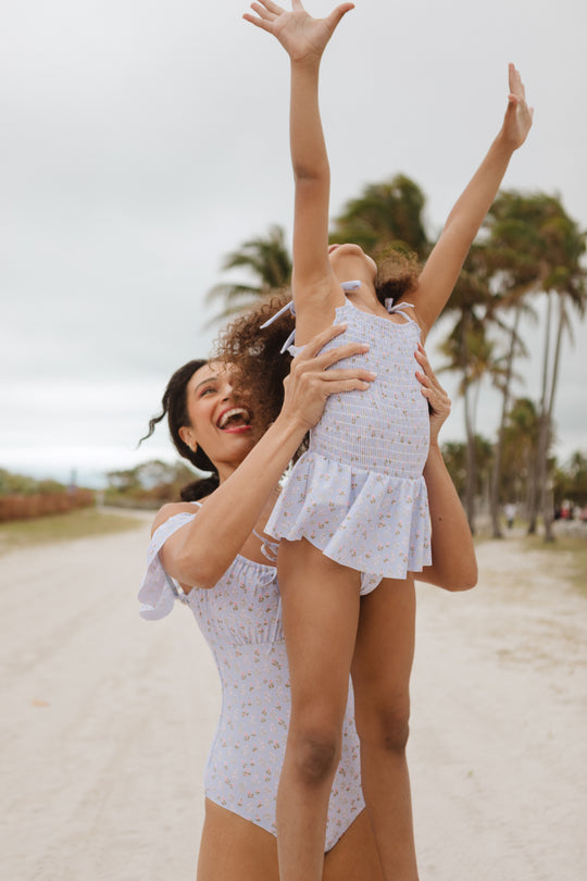 A woman in Ivy City Cos Mini Maggie One Piece in Blue Ditsy Floral lifts a laughing child, also in matching swimwear, into the air on a sandy beach with palm trees behind them.