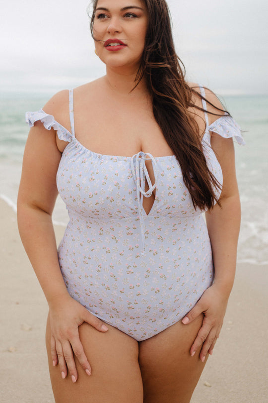 A woman stands on a sandy beach in the Ivy City Co Maise One Piece in Blue Ditsy Floral, featuring ruffled straps and a front tie, looking slightly away from the camera with the ocean behind her.