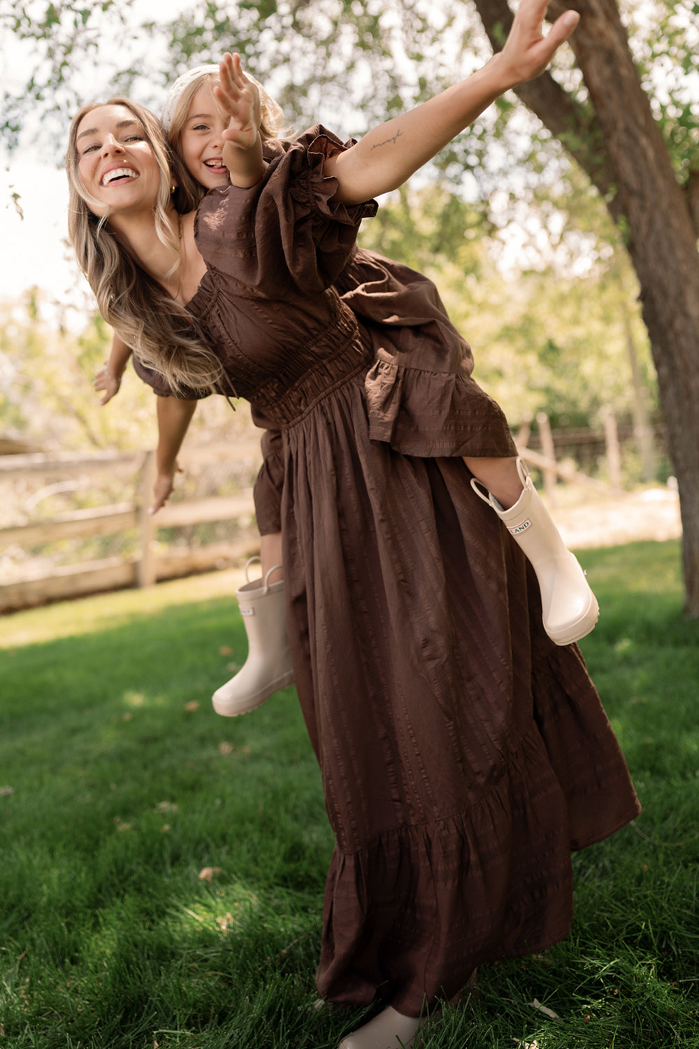 A smiling woman in the Ivy City Co Maleah Dress in Warm Cocoa gives a piggyback ride to a young girl wearing a matching dress and boots. With arms outstretched, they play together in a grassy, sunlit yard surrounded by trees and a wooden fence.