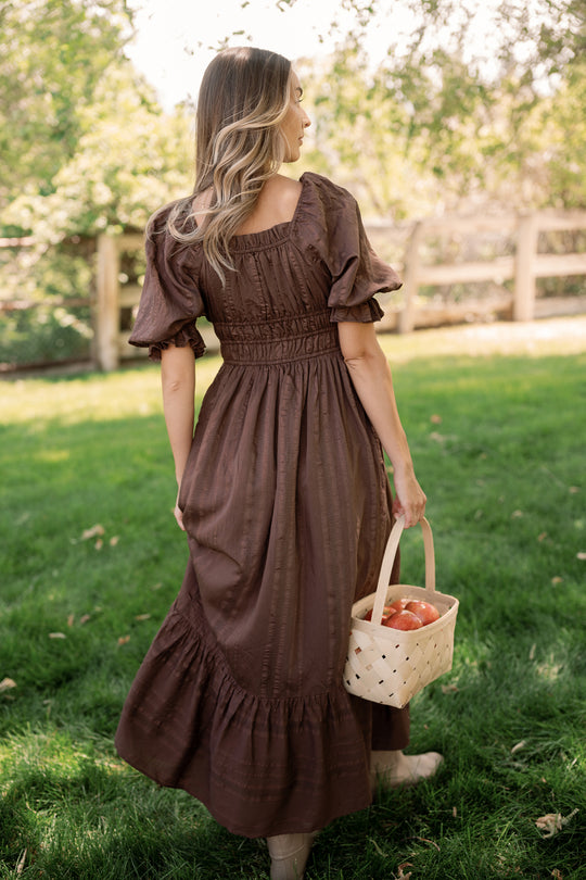 A woman wearing the Ivy City Co Maleah Dress in Warm Cocoa stands on green grass, holding a white basket of apples. She is facing away from the camera, with trees and a wooden fence in the background on a sunny day.