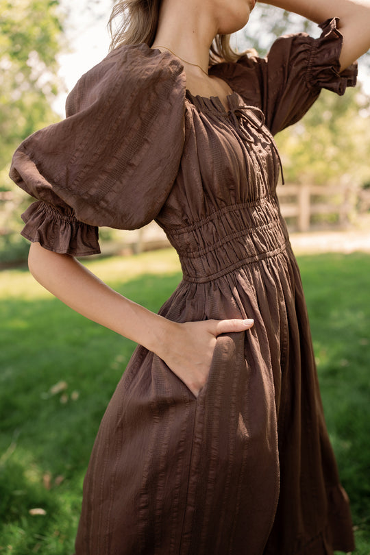 A woman stands outdoors on the grass, hand in pocket, wearing the Ivy City Co Maleah Dress in Warm Cocoa. The background is blurred with green trees and a wooden fence, and her face is partially out of frame.