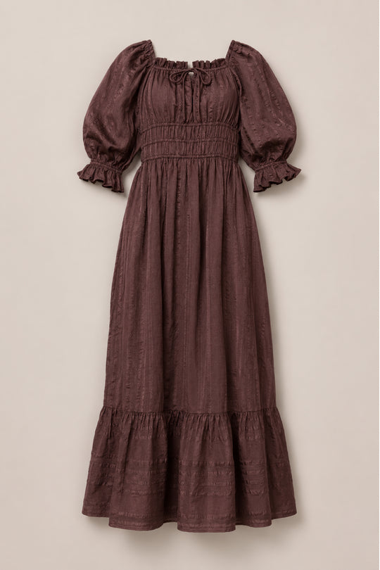 The Ivy City Co Maleah Dress in Warm Cocoa features a long maxi silhouette with puffed sleeves, ruffled cuffs, a smocked bodice, tie detail at the neckline, and a ruffled hem. Laid flat on a neutral background, this nursing-friendly style offers both comfort and elegance.